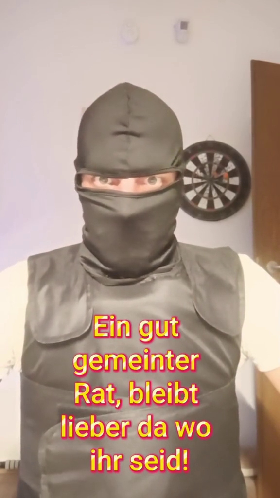Stephan M. schaut frontal in die Kamera und trägt dabei eine schusssichere Weste sowie eine Sturmhaube, Darüber Text: "Ein gut gemeinter Rat, bleibt lieber da wo ihr seid!"