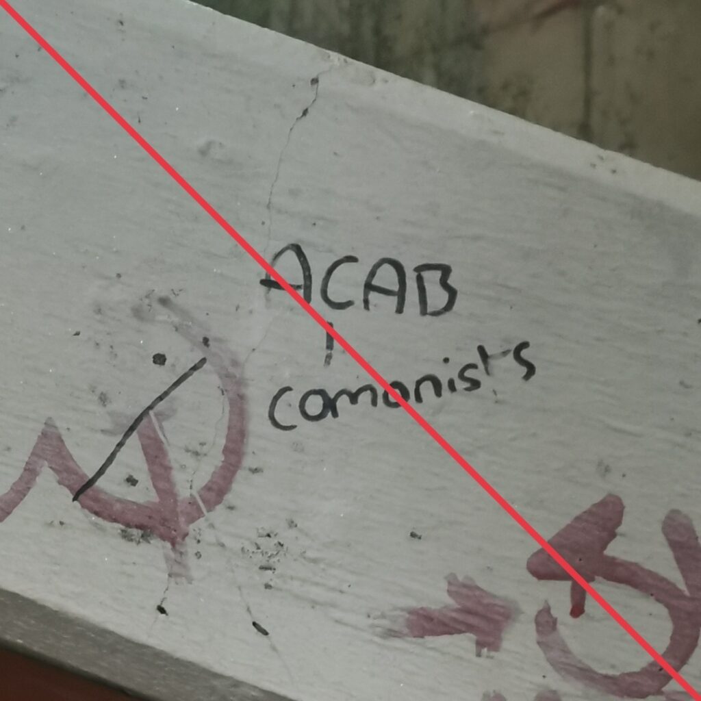 In schwarzem Marker geschriebener Schriftzug "ACAB" mit einer Linie, die von dem C auf das Wort "comonists" weist, neben einem durchgestrichenen Hammer und Sichel-Symbol, an einem Betonteil eines Treppenaufganges.