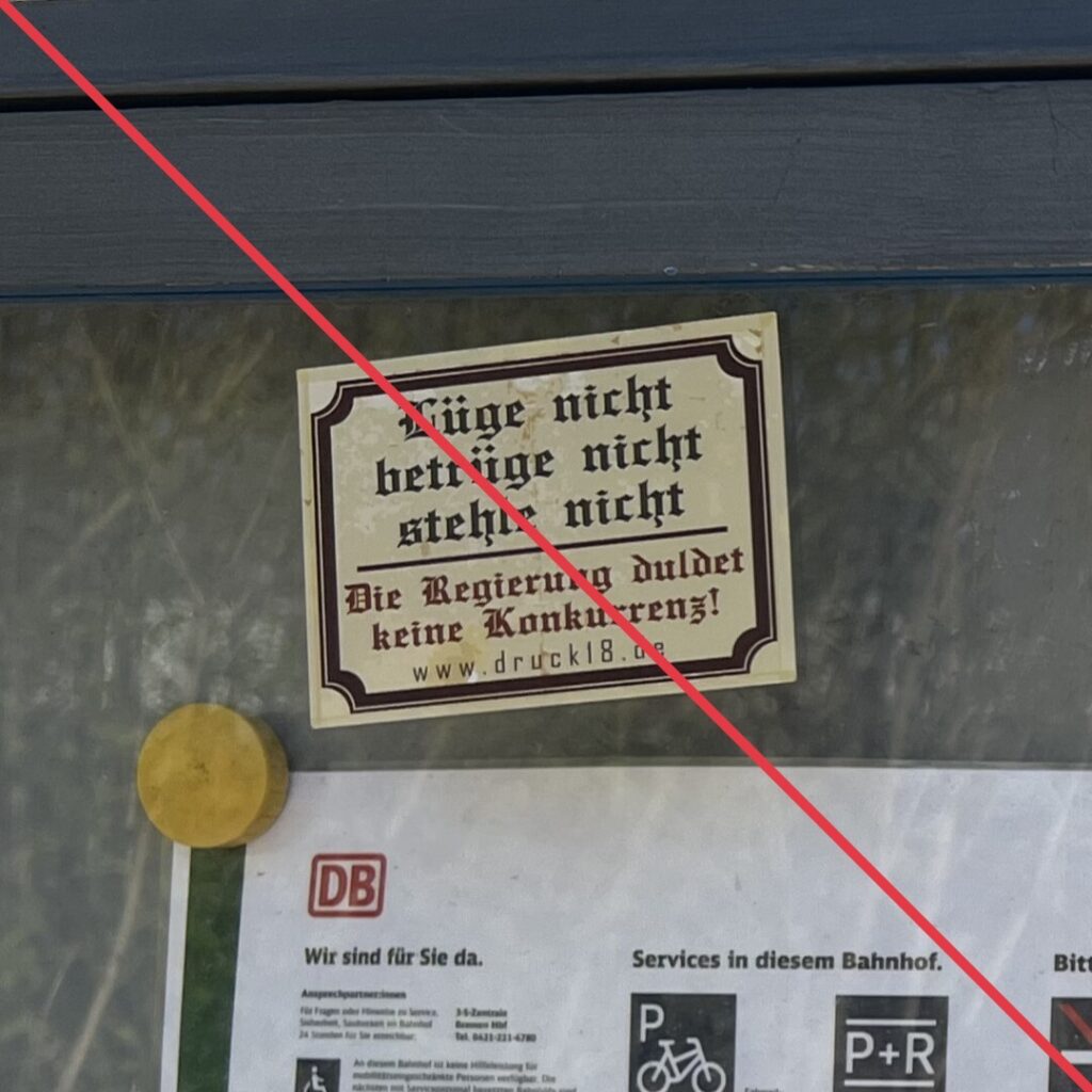 Aufkleber mit Aufschrift "Lüge nicht, betrüge nicht, stehle nicht - Die Regierung kennt duldet keine Konkurrenz!" auf der Scheibe einer Informationstafel.