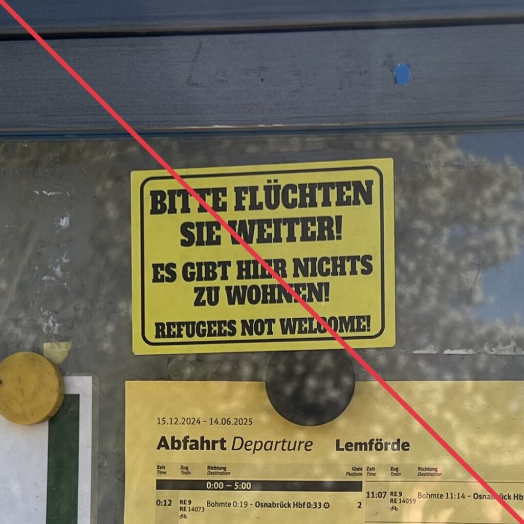 Aufkleber mit Aufschrift "Bitte flüchten Sie weiter! Es gibt hier nichts zu wohnen! Refugees not welcome!" auf der Scheibe einer Informationstafel.