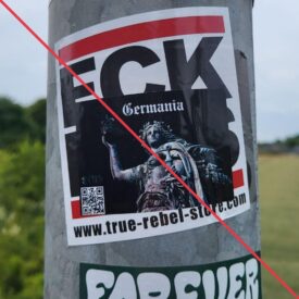 Aufkleber mit Aufschrift "Germania" über einer Abbildung der Germania des Niederwalddenkmalsund runenartigen Logo "Reyra" über einem QR-Code, der einen antifaschistischen Aufkleber überdeckt.