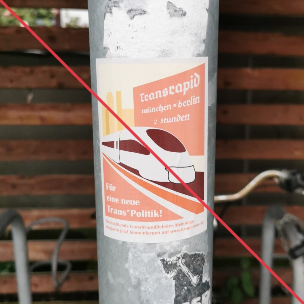 Aufkleber mit Aufschrift "Für eine neue Trans*Politik!" vor der Abbildung eines Transrapids und Werbung für eine extrem rechte Zeitschrift auf einem Mast eines Fahrradunterstands.