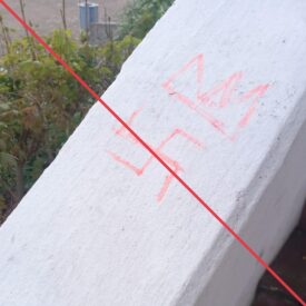Ein in rotem Stift gezeichnetes Hakenkreuz und darüber eine Krone, auf dem weißem Geländer einer Außentreppe.