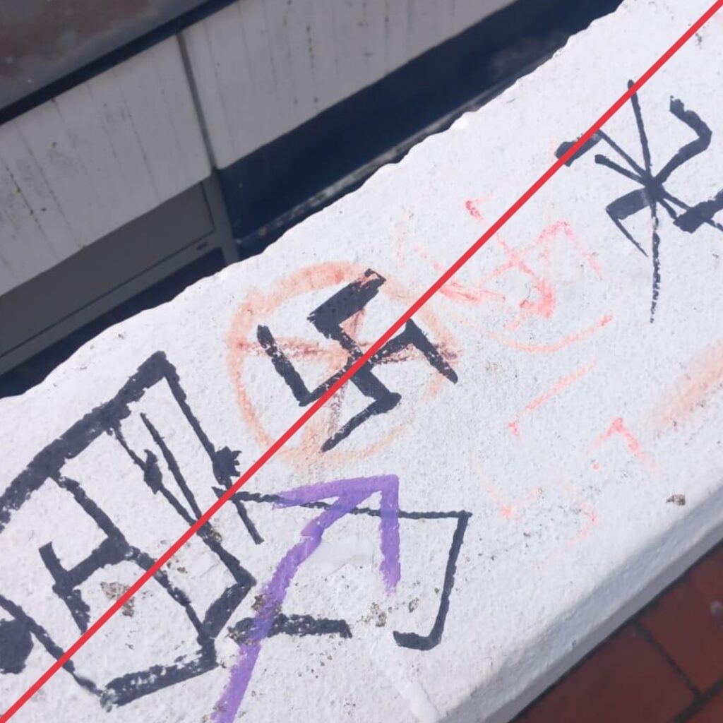 Mehrere Hakenkreuze, in schwarzem Marker und rotem Stift gezeichnet und teils durchgestrichen, auf dem weißen Geländer einer Außentreppe.